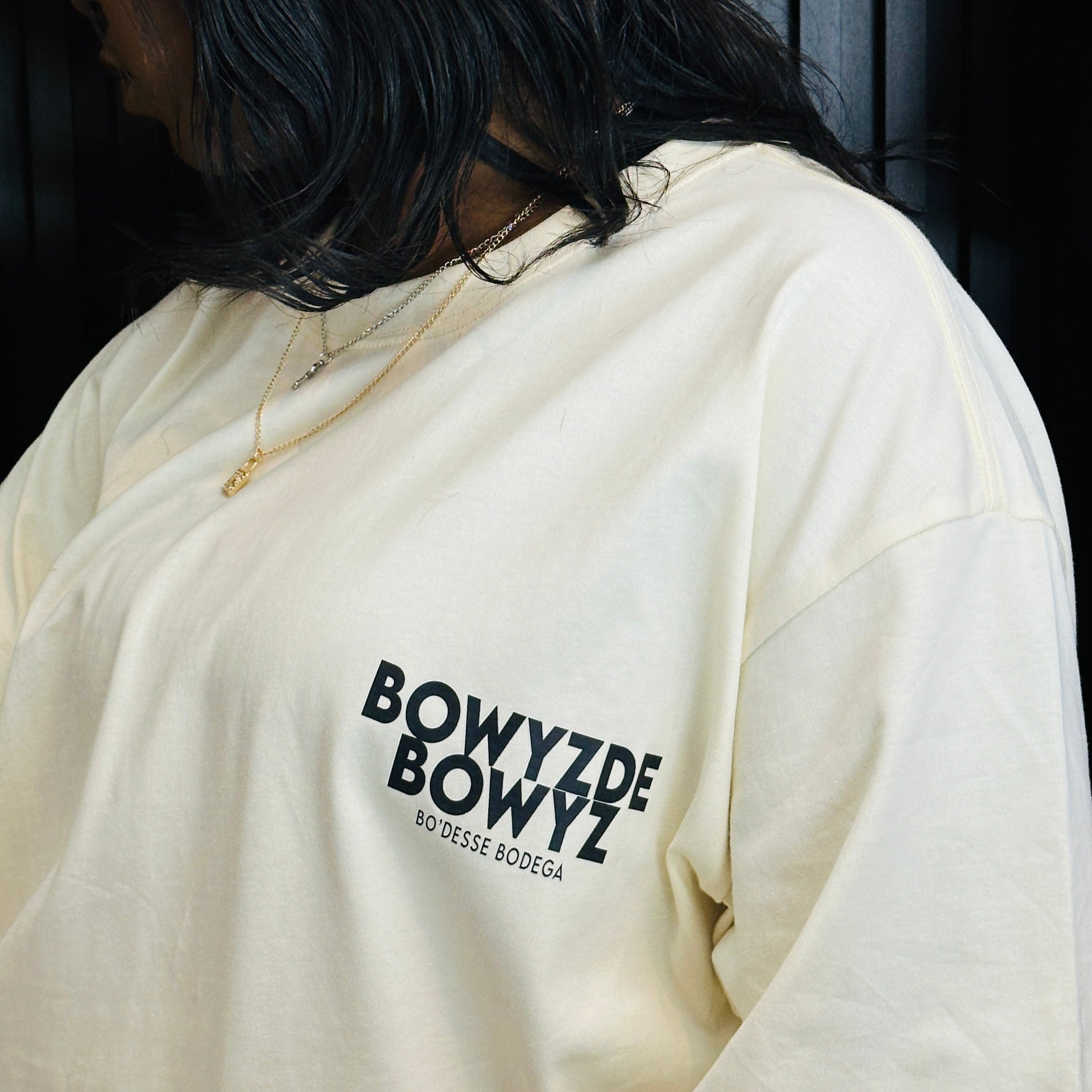 Person wearing a beige TEE with 'BOWYZ DE BOWYZ' text, standing against a dark background.